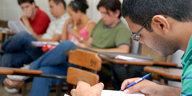 Fotografia de estudantes sentados em carteiras em uma sala de aula. Em destaque, um estudante observa uma prova segurando uma caneta.