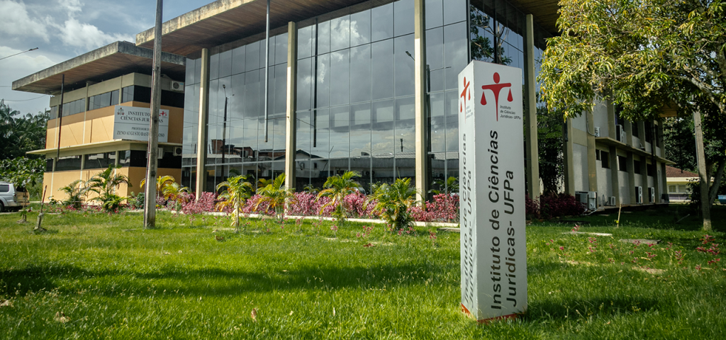 Fotografia da fachada do Instituto de Ciências Jurídicas da UFPA. Em frente, há um totem de identificação do prédio em um jardim com gramado.