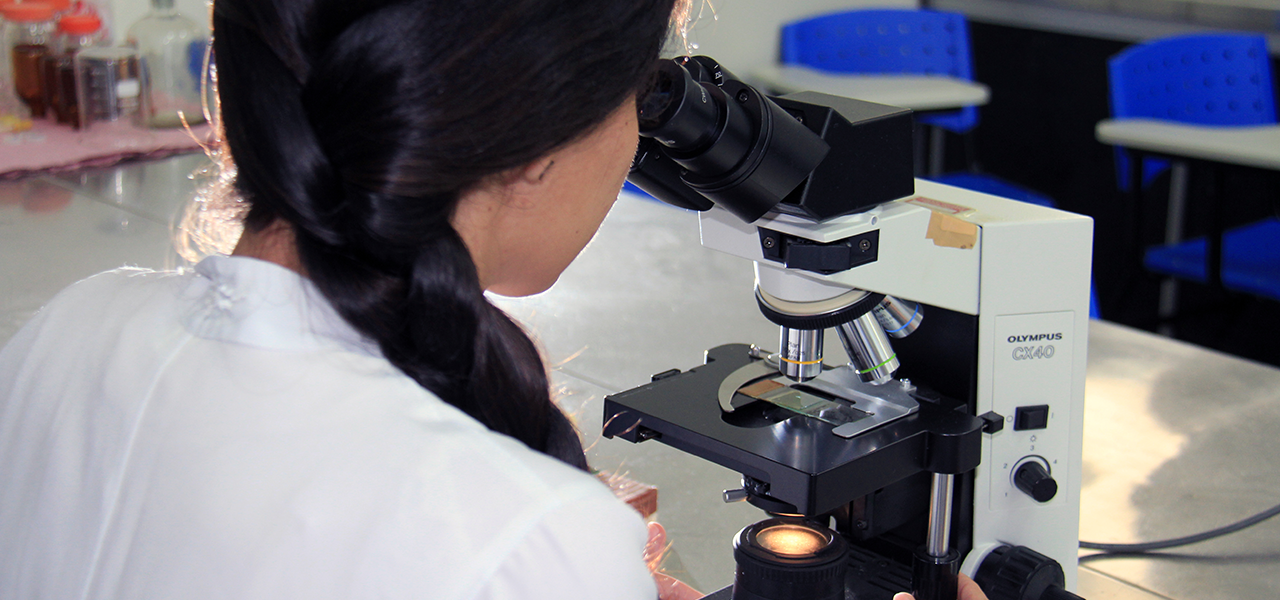 Fotografia de uma jovem de cabelos longos e pretos trançados, usando jaleco, observando uma amostra em um microscópio.