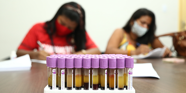 Fotografia de amostras de sangue em vidros de exames. Ao fundo, há duas mulheres sentadas escrevendo em um papel.