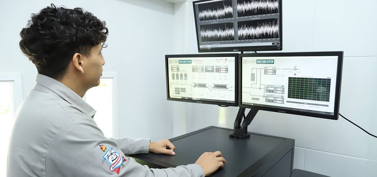 Fotografia de um estudante com jaleco cinza observando três telas de monitores com gráficos.