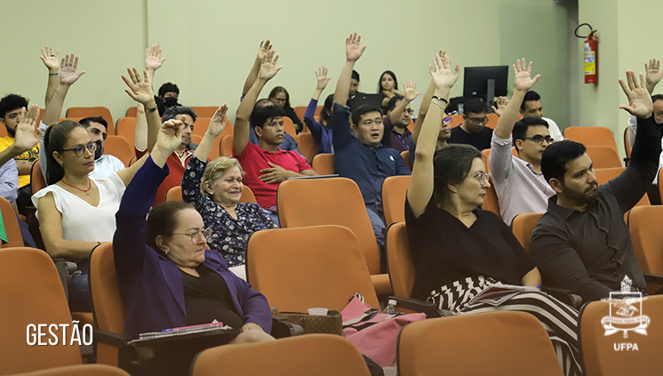 Fotografia de várias pessoas sentadas em um auditório com uma mão para cima participando de uma votação.