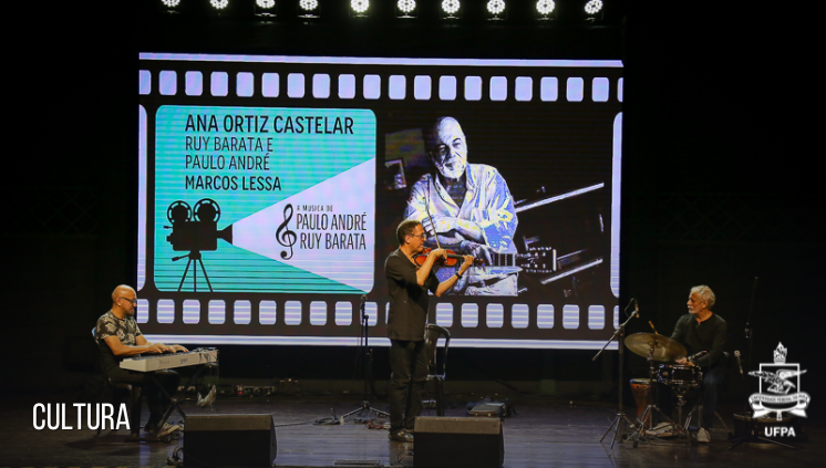 Fotografia de um trio musical sobre um palco. Os músicos tocam teclado, violino e percussão. Ao fundo, está um painel luminoso com a identidade visual do Álbum A Música de Paulo André e Ruy Barata e a foto de Paulo André com um violão.