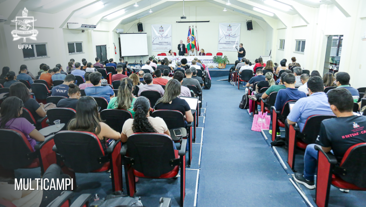 Fotografia registra as costas de uma plateia, num salão. Ao fundo, várias pessoas sentadas à mesa do evento.