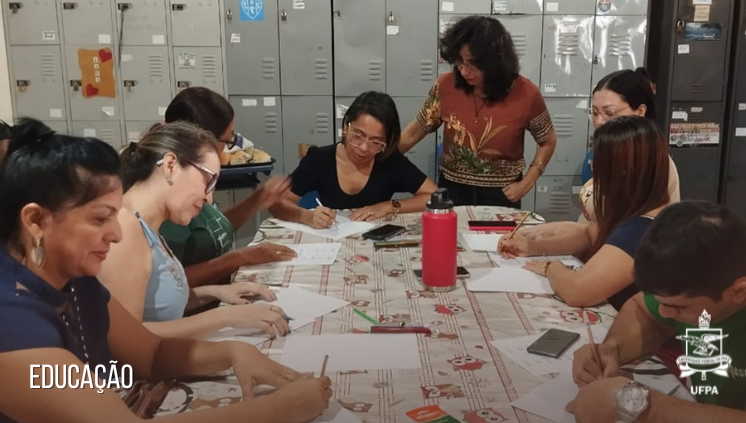 Fotografia com a imagem de um grupo de mulheres sentadas ao redor de uma mesa oval. As mulheres escrevem algo em folhas de papel disposta na mesa.