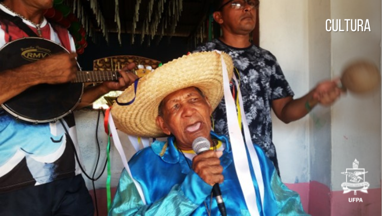 Fotografia de Zeferino Gonçalves dos Santos ( Mestre Zampa). Ele usa chapéu de palha, com fitas coloridas penduradas, e segura um microfone .
