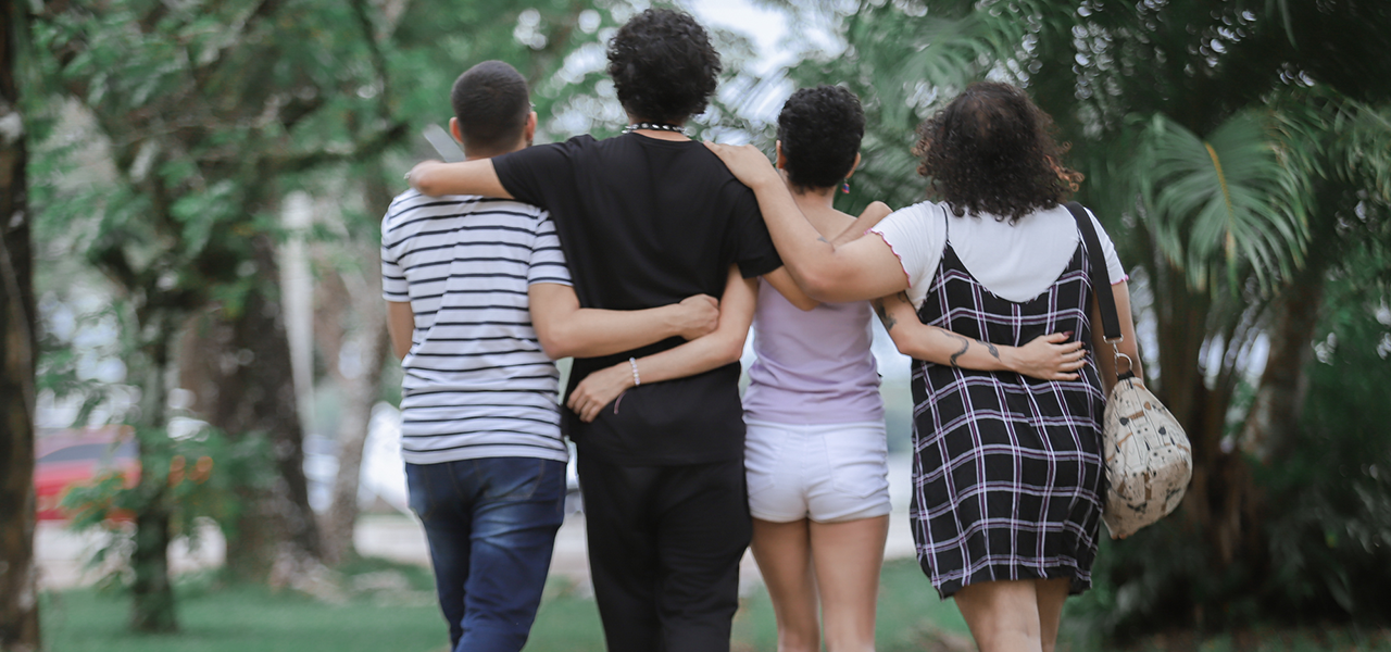 Fotografia de costas de quatro jovens caminhando abraçados em uma passarela arborizada.
