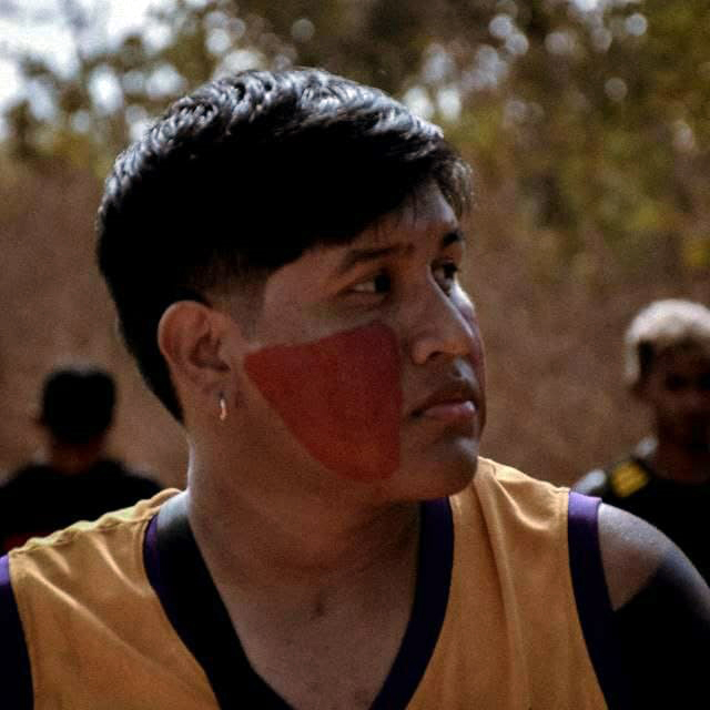 Foto do estudante indígena Riquelme Guara. Ele está de perfil e no rosto apresenta uma pintura indígena na cor vermelha.