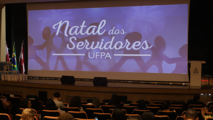Fotografia de um auditório com as luzes semiapagadas mostrando um telão no palco com uma arte gráfica com o texto "Natal dos Servidores da UFPA". Na plateia, as pessoas estão sentadas nas poltronas.