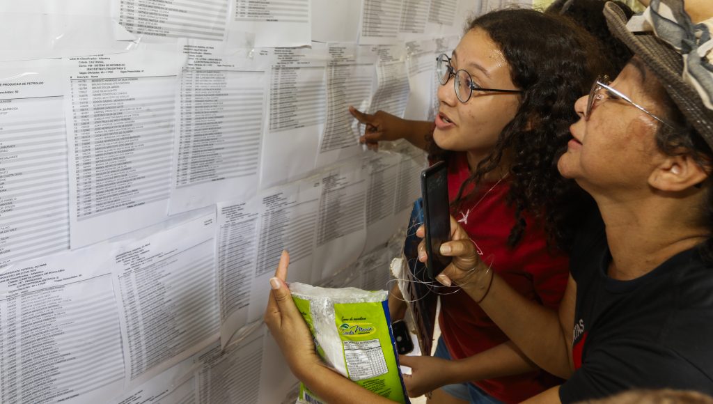 Duas pessoas estão atentas à folhas do listão de aprovados que estão dispostas em um painel. Em destaque na imagem, está a caloura Kaylanne Ribeiro.