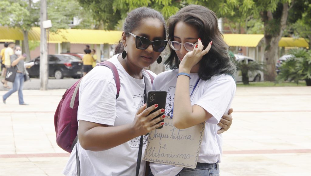 Fotografia de duas mulheres abraçadas, Renata e Bárbara Furtado. Elas olham para um aparelho celular. 