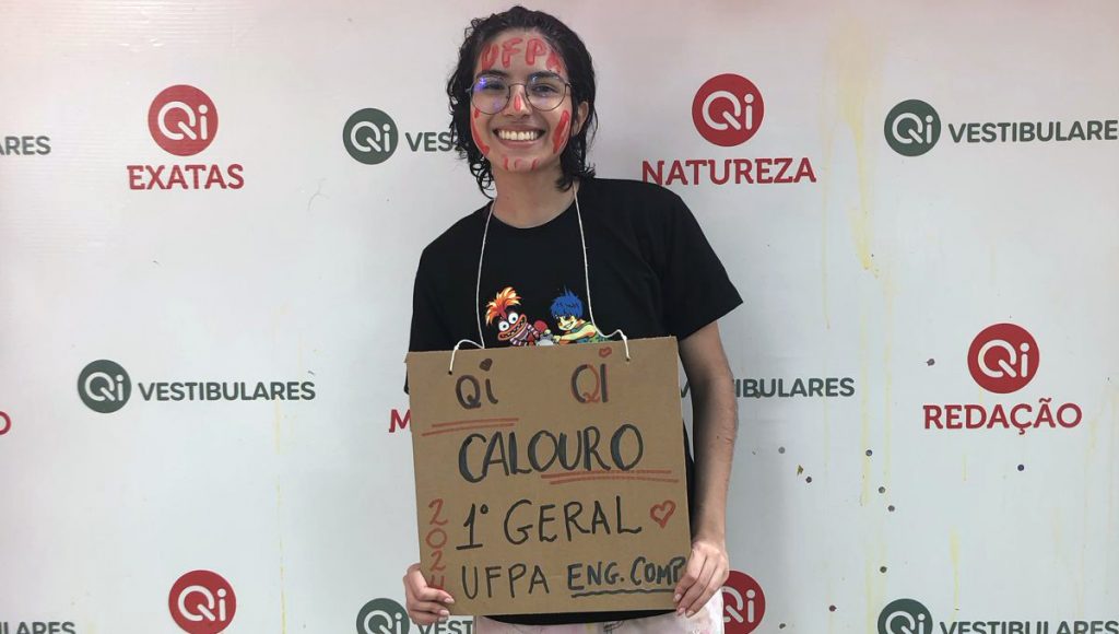Fotografia de uma menina jovem. Ela está sorrindo e segura uma placa com a mensagem: CALOURO PRIMEIRO LUGAR GERAL UFPA ENG. COMP.