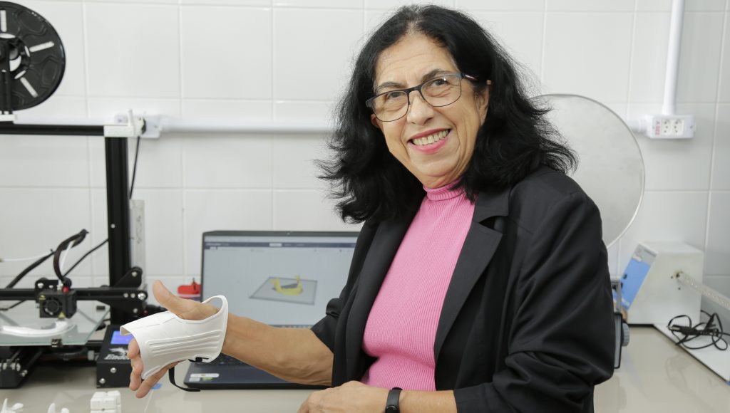Fotografia da Pró-reitora de Pesquisa e Pós-graduação da UFPA, professora Iracilda Sampaio, sorridente ao testar uma peça confeccionada na impressora 3D. A professora exibe a peça que está colocada em sua mão direita.