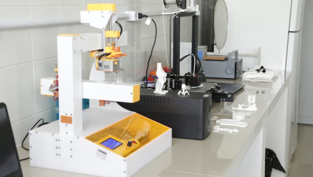 Fotografia de uma bancada do laboratório. Sobre esta bancada, diferentes tipos de equipamentos que serão utilizados no novo laboratório. e algumas peças já produzidas pela impressora 3D.