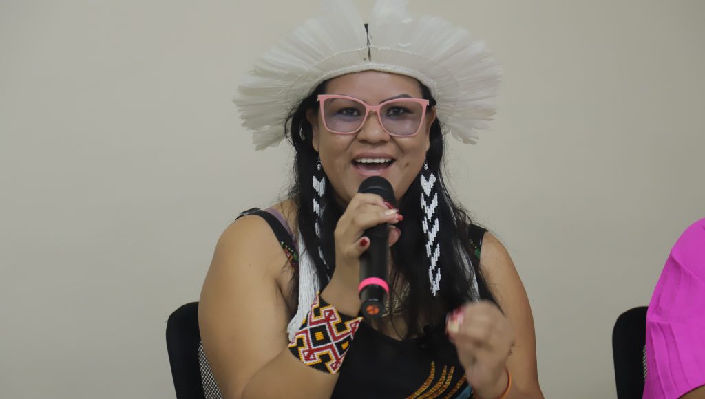 Fotografia de Josilene Nunes enquanto fala ao microfone. Josilene usa pulseira indígena no braço direito, brincos indígenas nas cores preto e branco, e um cocar na cor branca.