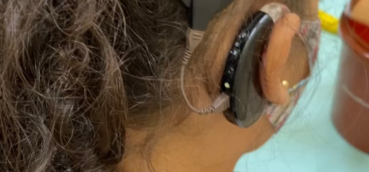 Fotografia de uma criança de costas. Ela está com o cabelo preso e usa um aparelho auditivo na orelha direita.