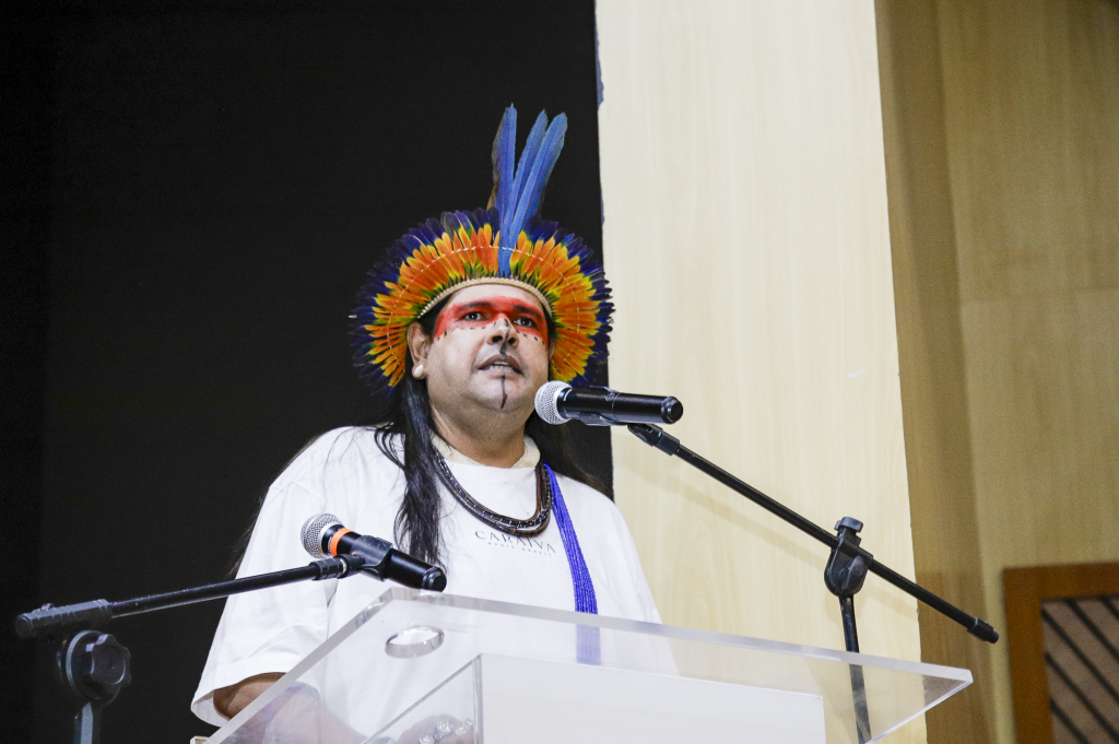 Fotografia do professor Uwira Xakriabá durante seu pronunciamento no púlpito. Ele usa camiseta branca lisa e acessórios de arte indígena, incluindo um cocar com penas de cor azul e amarela. Ele tem o rosto pintado na região dos olhos com tinta vermelha. 