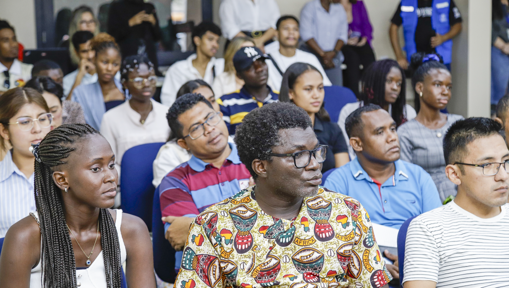 Fotografia destaca alguns dos estudantes presentes no evento, entre eles está o estudante angolano Scoth Cambolo. Scoth é um homem negro, usa camisa estampada com motivos africanos e óculos de armação na cor preta.