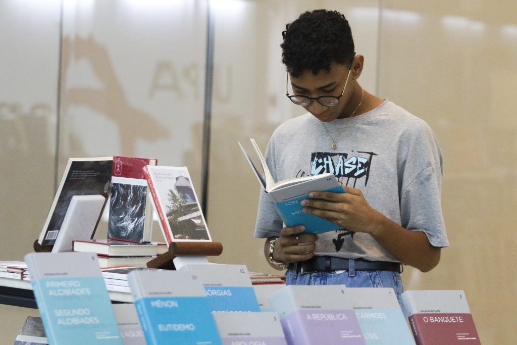 Fotografia de um jovem pardo lendo um livro. Ele está de pé, veste uma camiseta cinza e usa óculos. À sua frente há uma bancada com vários livros expostos. 