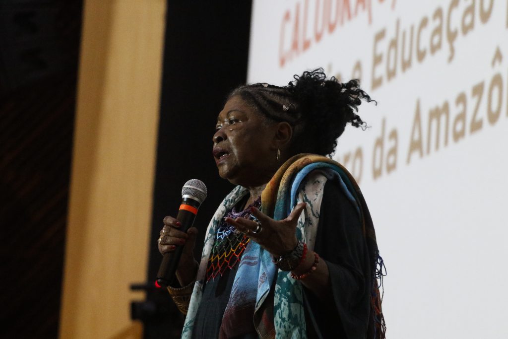 Fotografia da professora Zélia Amador de Deus, uma mulher negra que está com os cabelos trançados. Ela usa uma roupa escura com sobreposição de uma echarpe com estampa étnica. Zélia está falando ao microfone.   
