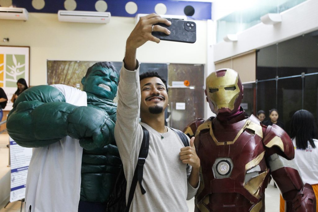 Fotografia de um jovem fazendo uma selfie ao lado das personagens Hulck e Homem de Ferro. O jovem está de mochila e faz um sinal de positivo com o dedo polegar.  