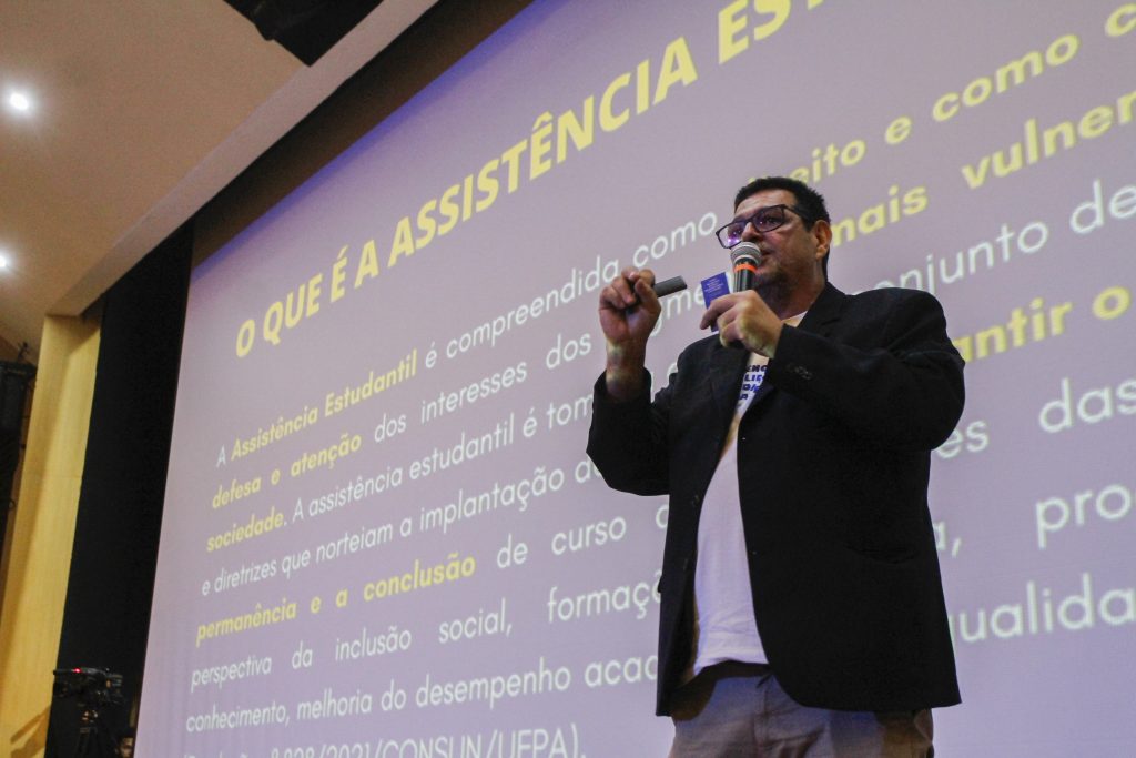 Fotografia do diretor da Saest, Ronaldo Araújo, um homem branco, de cabelos escuros e curtos. Ele está de pé, usa óculos e veste camisa de cor clara, blazer preto e calça jeans. Ele está falando ao microfone. 