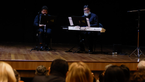 Fotografia de dois músicos performando em um palco com luz baixa. Um toca clarineta e outro, piano digital.