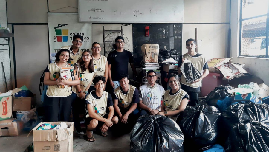 Fotografia posada de um grupo de voluntário do projeto Trote Solidário e Sustentável da UFPA. Eles usam colete da campanha na cor bege e estão em uma sala fechada rodeados por muitas caixas e sacos de doação.