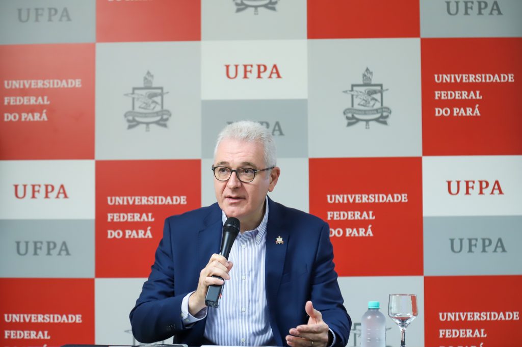 Fotografia do reitor da UFPA, Emmanuel Zagury Tourinho. Homem branco, grisalho e que usa óculos. Ele está sentado e fala ao microfone. Atrás dele, um banner com o brasão e a sigla UFPA repetida várias vezes. 