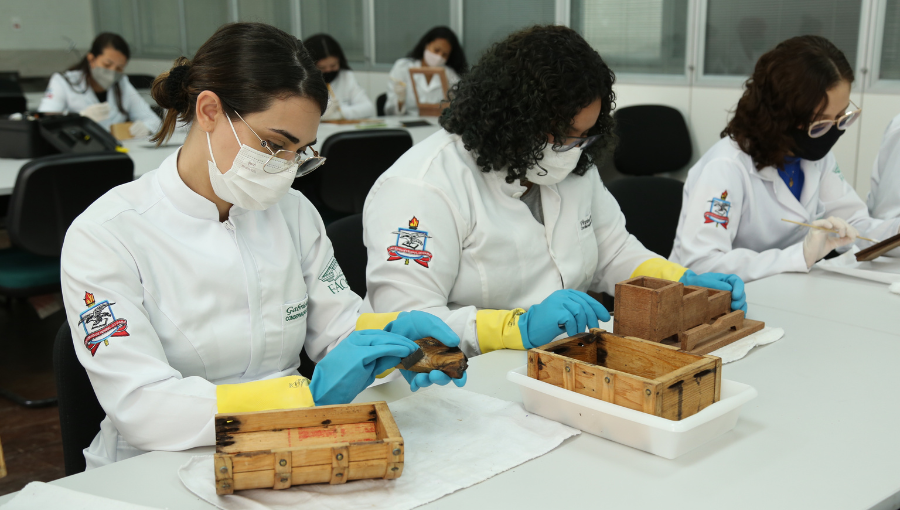 Fotografia evidencia três mulheres. Elas estão sentadas, usam jaleco branco com bordado do brasão da Universidade na manga direita. Todas fazem uso de luvas, máscara e óculos de grau. O trio manipula objetos de madeira sobre uma bancada de cor branca.