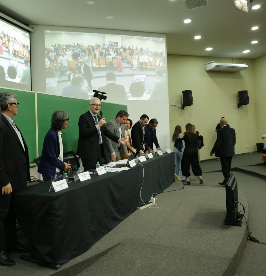 Fotografia da mesa onde estão reunidos os membros do Conselho Universitário representantes da Administração Superior. Todos estão de pé com destaque para o reitor Emmanuel Tourinho que fala ao microfone.