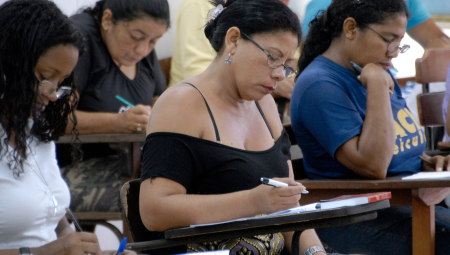 Fotografia mostra grupo de pessoas em sala de aula. Eles estão sentados e escrevendo sobre um papel individualmente.