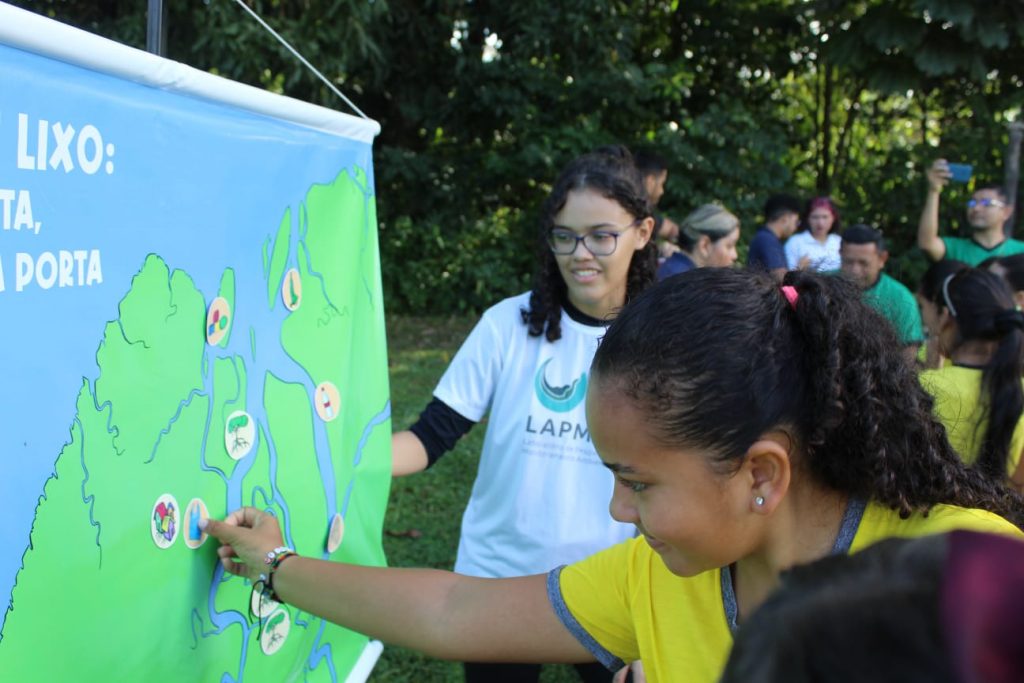 Fotografia demonstra uma menina de pele parda e cabelos cacheados colocando discos com figuras ecológicas em um mapa, enquanto uma mulher de camisa branca e óculos preto a auxilia na atividade do seu lado direito.