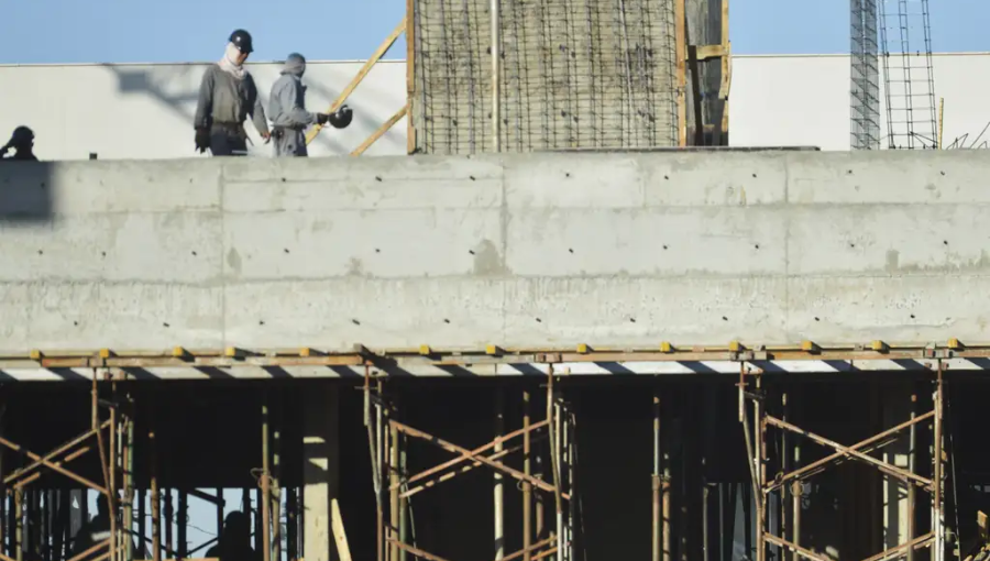 Fotografia mostra uma obra de construção civil. Na imagem, há estruturas de concreto, alicerces de apoio e dois operários usando equipamentos de proteção individual.