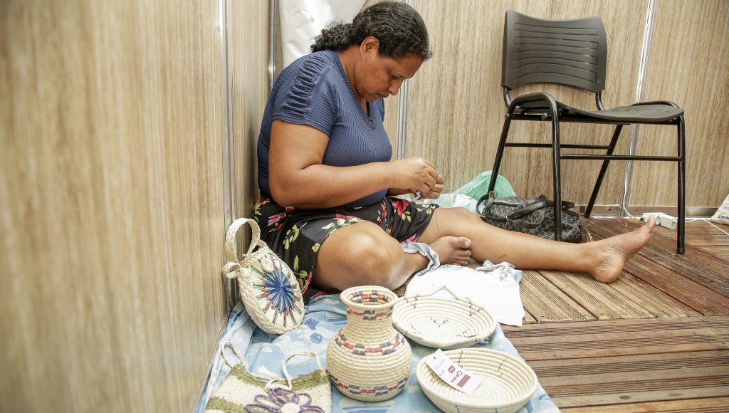 Fotografia de uma artesã tecendo uma peça. Ela está sentada no chão e ao seu redor estão dispostos algumas peças prontas, entre elas é possível observar: uma bolsa, um vaso, e dois pratos.