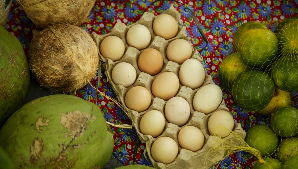 Fotografia de um cuba de ovos, alguns cocos verdes e secos e dois sacos de tangerina. Os alimentos estão disposto sobre um tecido florido nas cores vermelho, lilás e amarelo.