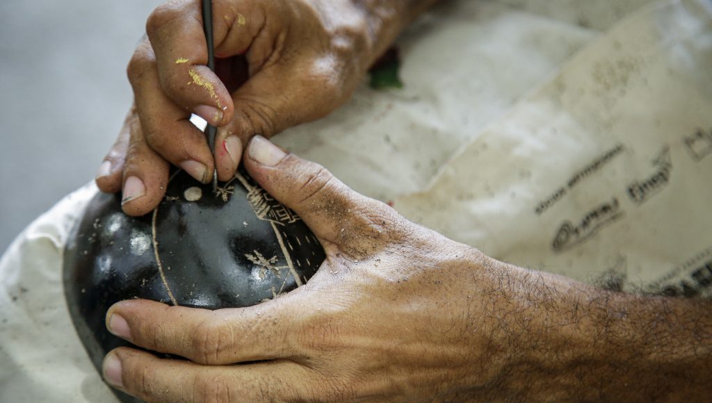 Fotografia das mãos de um artesão enquanto ele desenvolve um desenho em uma cuia preta.