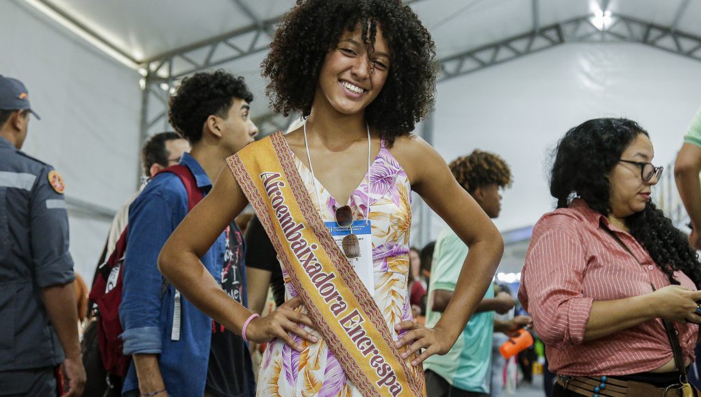 Fotografia de uma menina negra com cabelos cacheados na altura do ombro. Ela faz pose para a foto enquanto veste uma faixa com o seguinte texto: AFROEMBAIXADORA ENCRESPA.