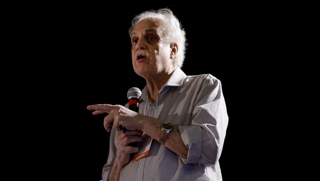 Fotografia do professor Carlos Nobre. Homem branco, com cabelos brancos, que veste uma camisa de manga comprida na cor cinza. Ele segura um microfone com a mão direita e gesticula com a esquerda.