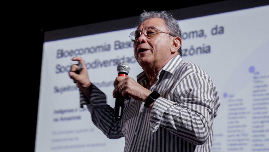 Fotografia do professor Francisco de Assis Costa. Ele fala ao microfone enquanto gesticula com a mão direta. O professor tem cabelos curtos e grisalhos e veste uma blusa de manga comprida nas cores branco e cinza.