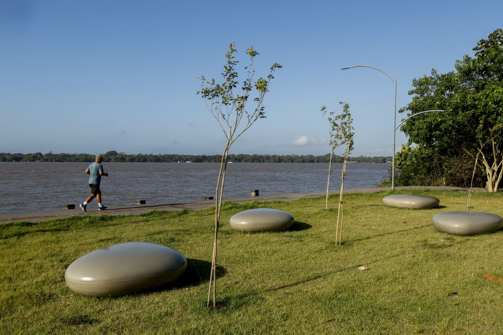 Fotografia de dia, próximo à orla do rio Guamá, em destaque, alguns bancos feitos no formato de pedra-sabão. 