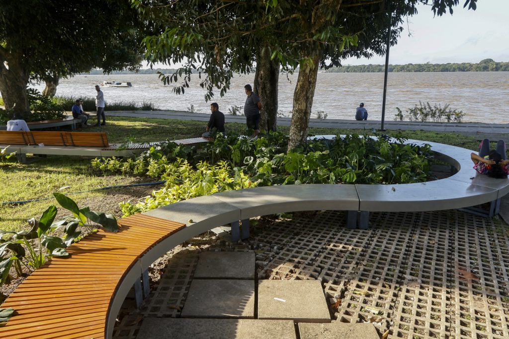 Fotografia de dia, próximo à orla do rio Guamá, em destaque, um banco comprido, em formato curvo, se estende em um jardim. Algumas pessoas estão sentadas no banco. 