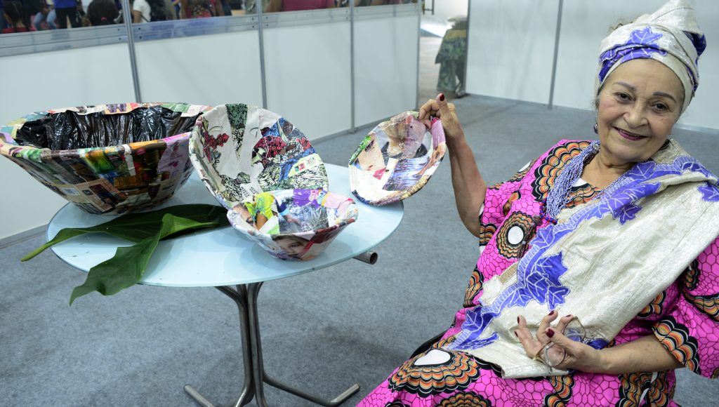 Fotografia de Mametu Nangetu. Ela usa um turbante nas cores branco e lilás e segura, com a mão direita, um alguidar produzido em papel. Ao seu lado há uma mesa com várias unidades de alguidar.
