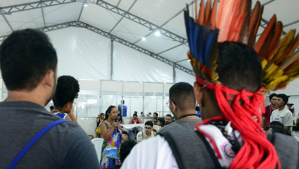 Fotografia interna da tenda Afro e Indígena. Um mulher, com pintura indígena no rosto, fala ao microfone, enquanto é observada por outras pessoas. Em destaque, no primeiro plano, a imagem de uma pessoa com cocar.