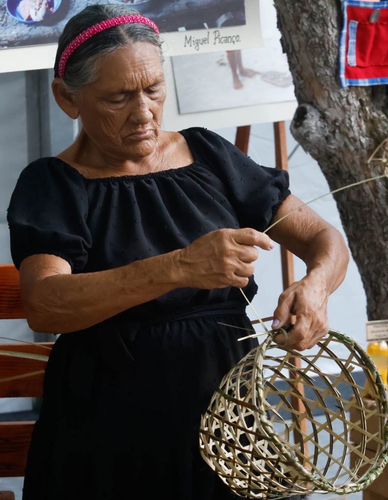 Fotografia uma senhora tecendo um paneiro de palha. Ela olha para o paneiro enquanto tece com as mãos. Ela tem cabelos grisalhos, usa uma tiara rosa e um vestido preto.
