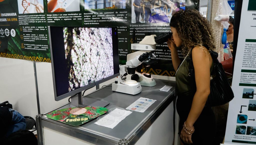 A imagem mostra uma mulher de cabelos cacheados e blusa verde olhando em um microscópio. O microscópio está sobre uma mesa e ao seu lado há uma tela mostrando uma imagem.
