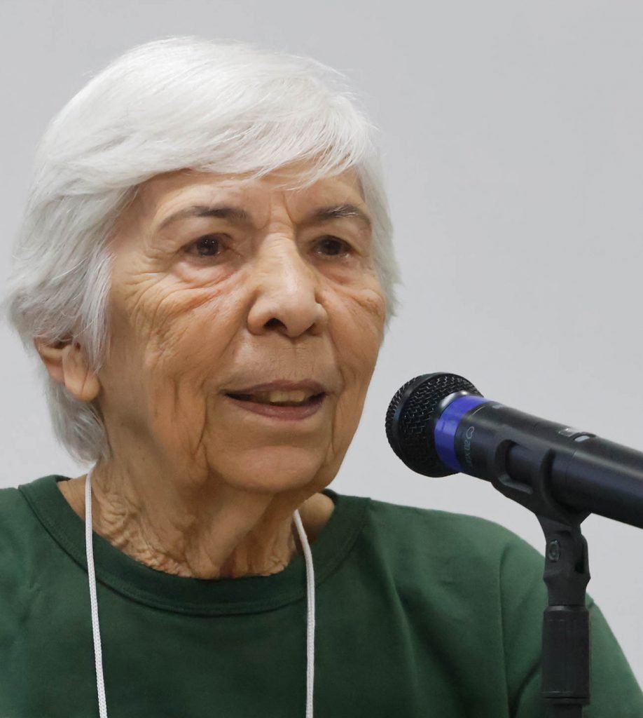 Fotografia close de Yvonne Mascarenhas. Uma mulher branca, de cabelos brancos, que veste uma blusa verde. A sua frente há um microfone preto.