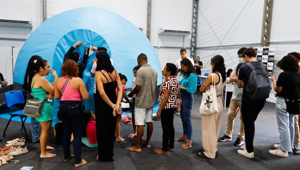Fotografia da parte interna de uma tenda. No primeiro plano, há uma fila com diversas pessoas. No fundo, um circulo azul inflável que imita a esfera de um planeta.
