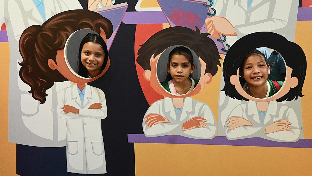 Fotografia de uma arte que reproduz as vestimentas de pequenos cientistas. Na arte há um círculo para que as pessoas possam colocar seus rostos. Três meninas crianças estão colocadas nesses círculos posando para a foto.