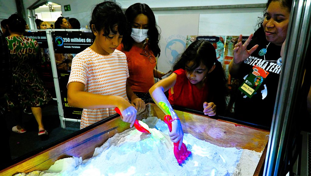 Fotografia de duas jovens meninas mexendo em uma caixa de areia com garfos plásticos de brinquedo. Elas são observadas por duas mulheres adultas.